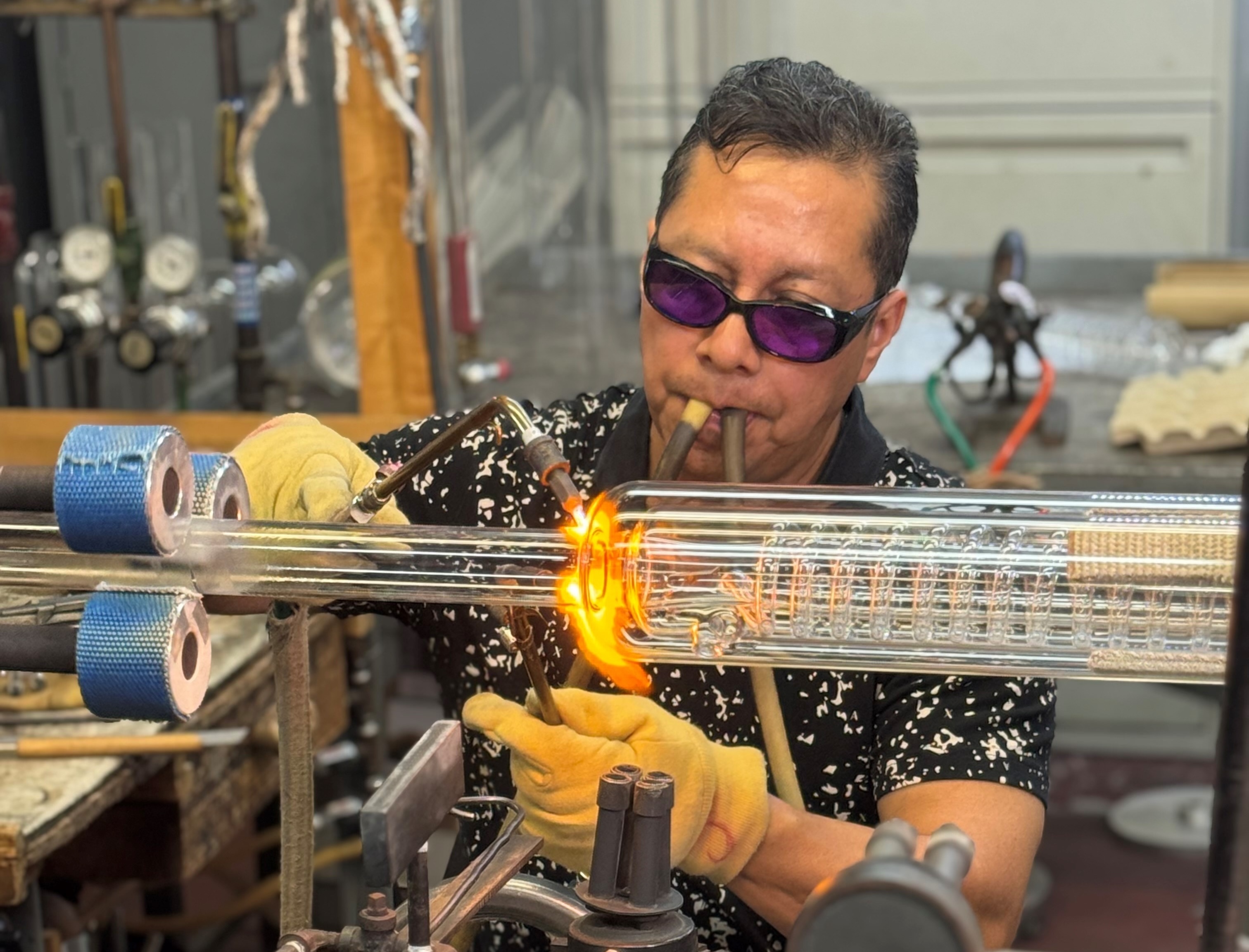 A man heating a glass apparatus with a blow torch. He has goggles on and two tubes in his mouth.