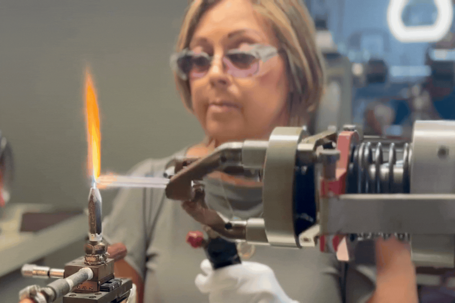 A machine turns a small glass tube while a woman aims a blowtorch at it.